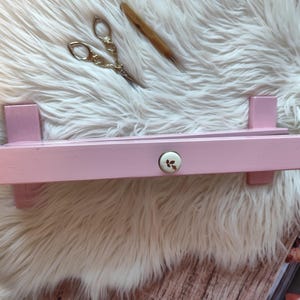 May include: A light pink wooden shelf with a decorative floral knob. The shelf is rectangular with two vertical supports on each end. A pair of gold scissors and a wooden tool rest on a white fluffy surface.