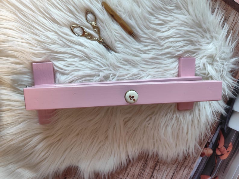 May include: A light pink wooden shelf with a decorative floral knob. The shelf is rectangular with two vertical supports on each end. A pair of gold scissors and a wooden tool rest on a white fluffy surface.