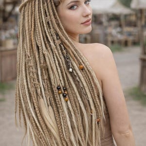 Puede incluir: Una mujer con largas rastas rubias, adornadas con cuentas y elementos decorativos. Las rastas caen por la espalda, con algunos mechones trenzados. La imagen está tomada al aire libre, con un fondo borroso.