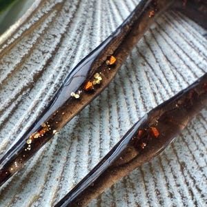 May include: Two brown hair clips with gold and orange glitter embedded in the resin. The clips are on a textured surface.