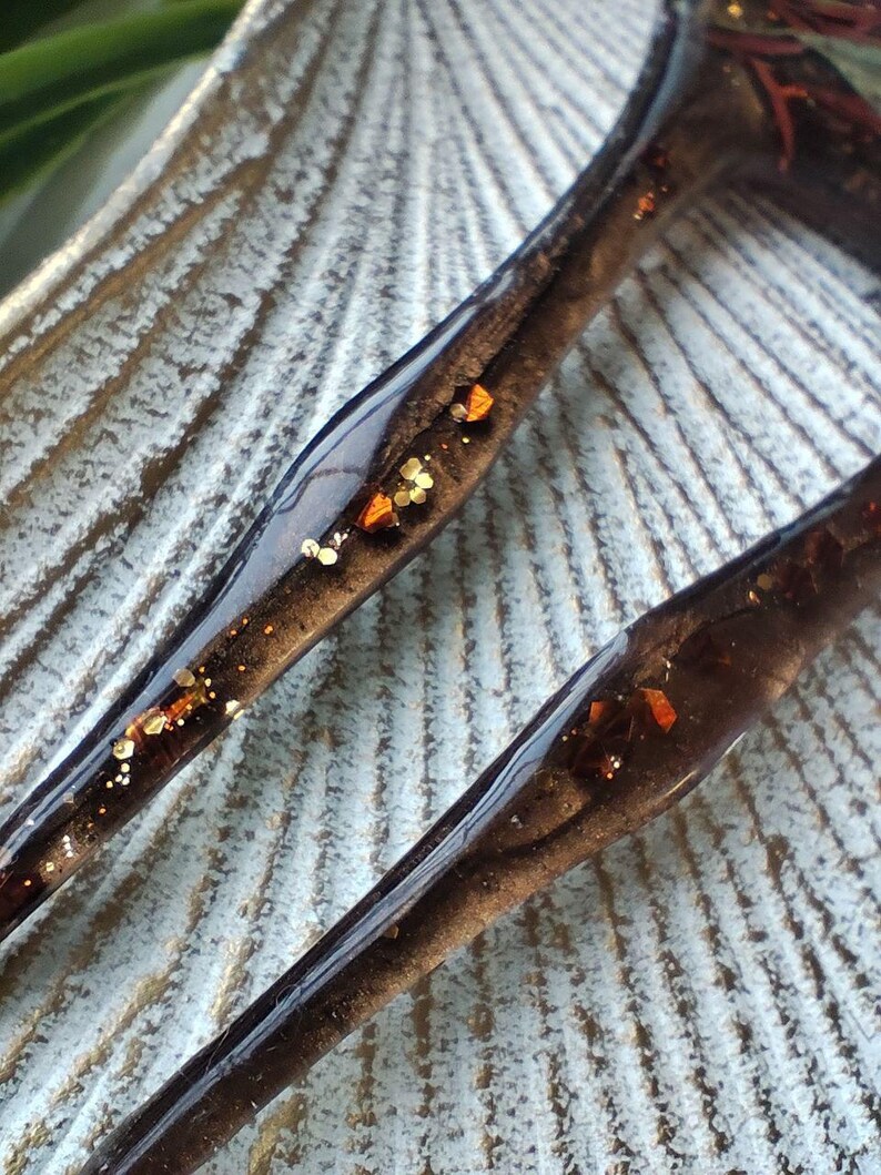 May include: Two brown hair clips with gold and orange glitter embedded in the resin. The clips are on a textured surface.