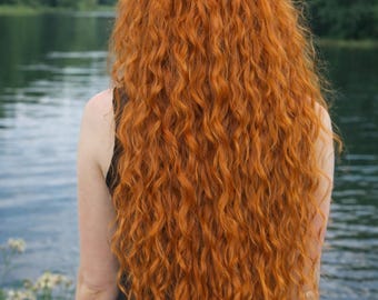 Op de afbeelding: Achteraanzicht van een persoon met lang, levendig rood krullend haar. Een zilveren haaraccessoire met groene edelstenen siert het haar. De persoon draagt een zwarte top, met een meer en bomen op de achtergrond.