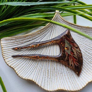 May include: A brown hair fork with a unique design featuring embedded dried flowers and gold glitter. The fork is resting on a white ceramic dish.