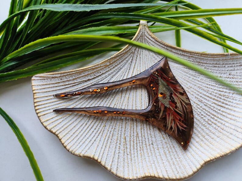 May include: A brown hair fork with a unique design featuring embedded dried flowers and gold glitter. The fork is resting on a white ceramic dish.