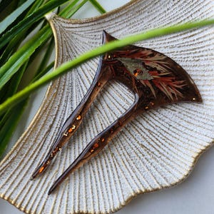 May include: A brown hair comb with a decorative design of red and gold glitter embedded in clear resin. The comb has two prongs and is displayed on a white and gold decorative plate.