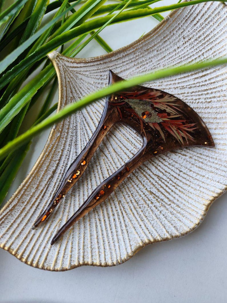 May include: A brown hair comb with a decorative design of red and gold glitter embedded in clear resin. The comb has two prongs and is displayed on a white and gold decorative plate.