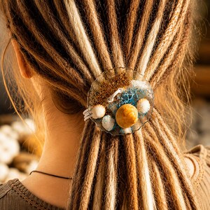 May include: A circular hair accessory featuring a clear resin base embedded with gold glitter, blue pigment, and three small natural seashells. The piece is secured with a white elastic band, designed to hold a section of hair in place with its textured edge.