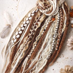 Op de afbeelding: Handgemaakte dreadlocks in bruin, beige en wit. De dreads zijn versierd met decoratieve stiksels, kralen en witte pompons. De opstelling is kunstzinnig vormgegeven, waarbij de ingewikkelde details van elke streng worden getoond.