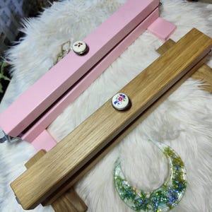 May include: Two wooden display stands, one pink and one natural wood, each with a decorative floral button. A crescent-shaped resin piece with green and blue inclusions rests nearby. The stands are designed to hold items for display.