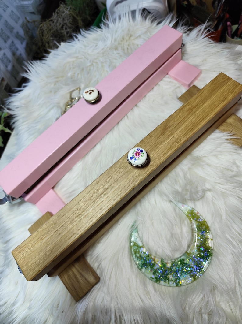 May include: Two wooden display stands, one pink and one natural wood, each with a decorative floral button. A crescent-shaped resin piece with green and blue inclusions rests nearby. The stands are designed to hold items for display.
