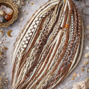 Op de afbeelding: Handgemaakte dreadlocks in bruin, beige en wit. De dreads hebben ingewikkelde patronen, waaronder geweven en gevlochten secties, met decoratieve kralen en accenten. De opstelling wordt gepresenteerd op een neutrale ondergrond.