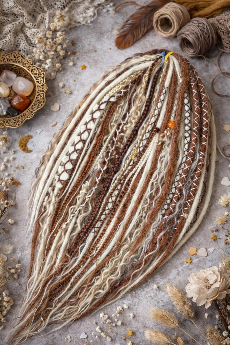 Op de afbeelding: Handgemaakte dreadlocks in bruin, beige en wit. De dreads hebben ingewikkelde patronen, waaronder geweven en gevlochten secties, met decoratieve kralen en accenten. De opstelling wordt gepresenteerd op een neutrale ondergrond.
