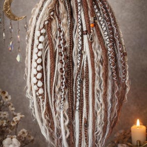 Op de afbeelding: Een verzameling bruine, witte en beige dreadlocks, sommige met decoratieve kralen en stiksels. De dreadlocks zijn gestyled met een gele band aan de bovenkant. De afbeelding heeft een warme, aardse toon, wat duidt op een bohemien of alternatieve esthetiek.
