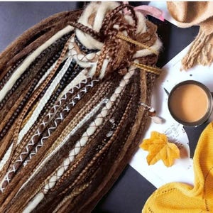 K&ouml;nnte beinhalten: Nahaufnahme eines B&uuml;ndels brauner, beiger und wei&szlig;er synthetischer Dreadlocks mit braunen N&auml;hten. Die Dreadlocks sind f&auml;cherf&ouml;rmig angeordnet und liegen auf einer wei&szlig;en Oberfl&auml;che.