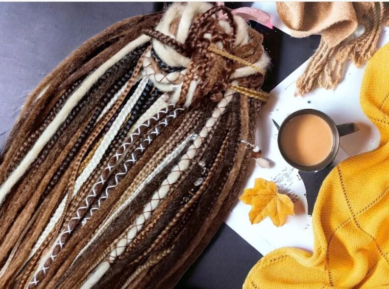 K&ouml;nnte beinhalten: Nahaufnahme eines B&uuml;ndels brauner, beiger und wei&szlig;er synthetischer Dreadlocks mit braunen N&auml;hten. Die Dreadlocks sind f&auml;cherf&ouml;rmig angeordnet und liegen auf einer wei&szlig;en Oberfl&auml;che.