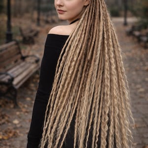 Puede incluir: Una mujer con largas rastas rubias, peinadas en un estilo recogido a medias. Lleva un top negro con los hombros descubiertos y mira por encima del hombro. El fondo es una escena borrosa al aire libre con bancos y árboles.