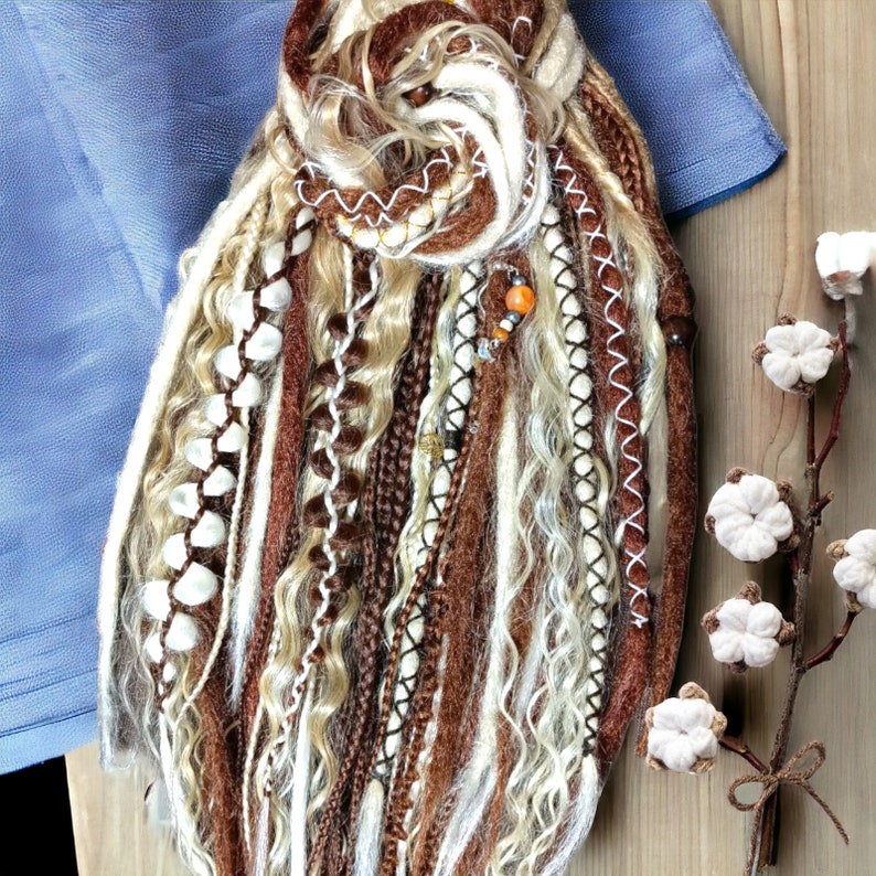 Op de afbeelding: Een verzameling bruine, cr&egrave;me en blonde dreadlocks, sommige met witte stiksels en decoratieve kralen. De dreadlocks zijn gerangschikt in een bundel, met een blauwe stof op de achtergrond. Een takje katoenbloemen staat op de voorgrond.