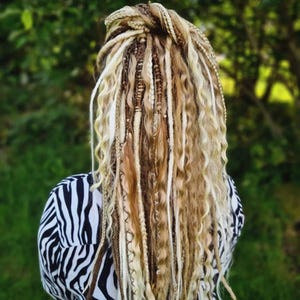 May include: A person with long, blonde, and brown dreadlocks with beads woven in. The dreadlocks are pulled back into a ponytail. The person is wearing a black and white zebra print shirt.