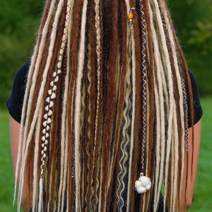Op de afbeelding: Close-up van lange, veelkleurige dreadlocks. Het haar heeft een mix van bruine, blonde en lichtbruine lokken, met decoratieve kralen en bedels. De dreadlocks vallen naar beneden en cre&euml;ren een getextureerd en gelaagd effect.