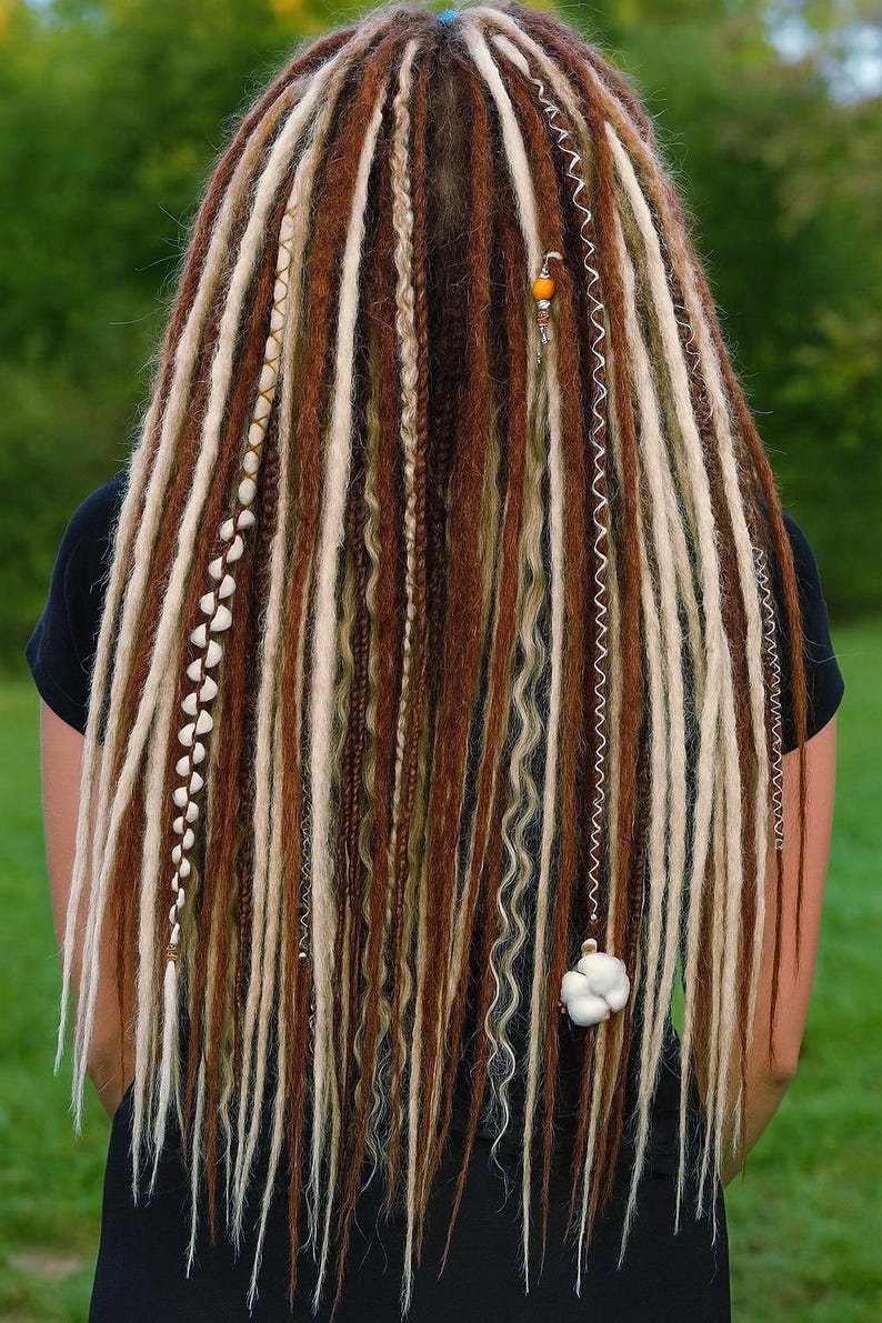 Op de afbeelding: Close-up van lange, veelkleurige dreadlocks. Het haar heeft een mix van bruine, blonde en lichtbruine lokken, met decoratieve kralen en bedels. De dreadlocks vallen naar beneden en cre&euml;ren een getextureerd en gelaagd effect.