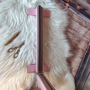 May include: A pink wooden frame with a dark gray interior, possibly a mirror or picture frame, is displayed on a white fluffy surface. A small pair of gold scissors and a wooden tool are also visible. The frame is hinged and appears to be foldable.