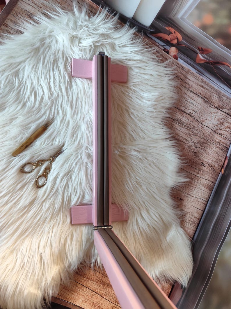 May include: A pink wooden frame with a dark gray interior, possibly a mirror or picture frame, is displayed on a white fluffy surface. A small pair of gold scissors and a wooden tool are also visible. The frame is hinged and appears to be foldable.