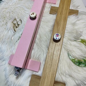 May include: Two wooden embroidery hoops, one painted pink and the other natural wood. Each hoop has a decorative knob with a floral design. The hoops are designed to hold fabric taut for embroidery or cross-stitch projects.