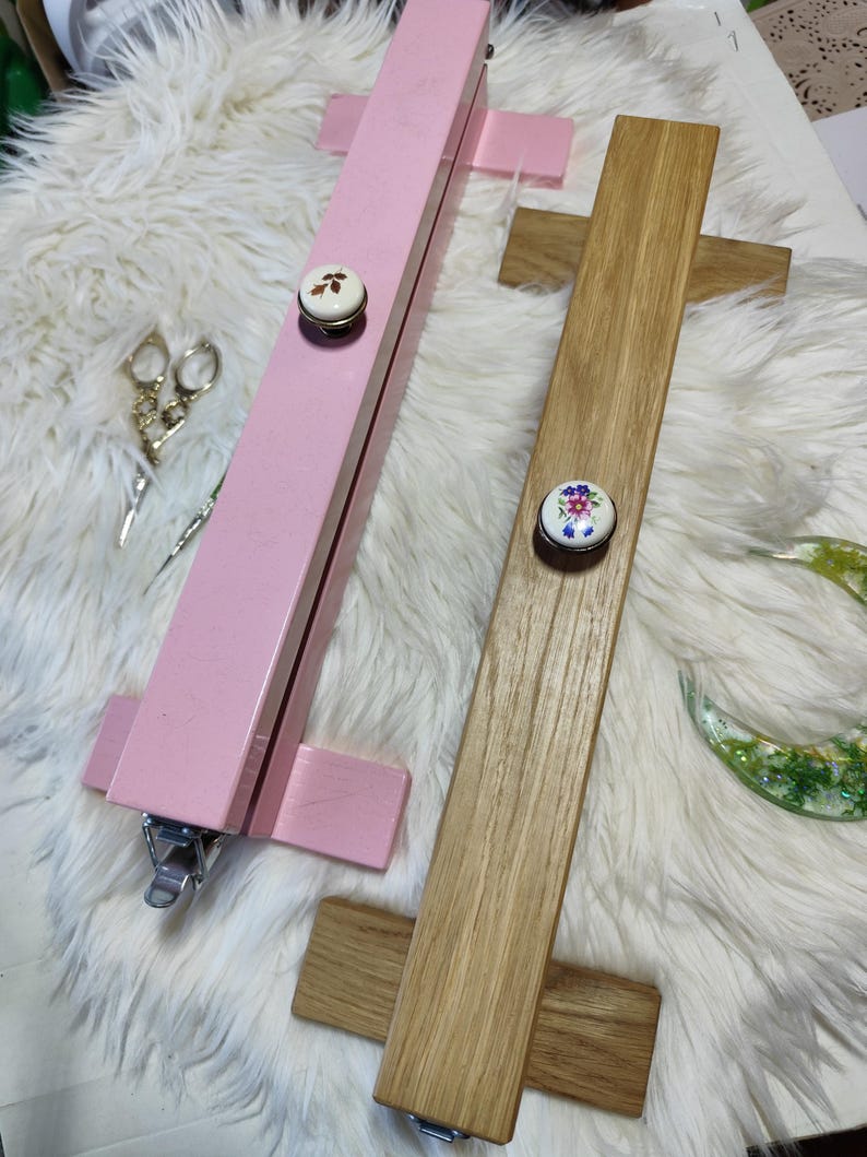 May include: Two wooden embroidery hoops, one painted pink and the other natural wood. Each hoop has a decorative knob with a floral design. The hoops are designed to hold fabric taut for embroidery or cross-stitch projects.