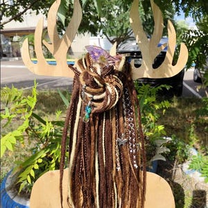 K&ouml;nnte beinhalten: Braune und wei&szlig;e synthetische Dreadlocks mit Perlen und Charms, die daran h&auml;ngen. Die Dreadlocks sind zu einem Knoten gebunden und an einem h&ouml;lzernen Hirschkopf befestigt.