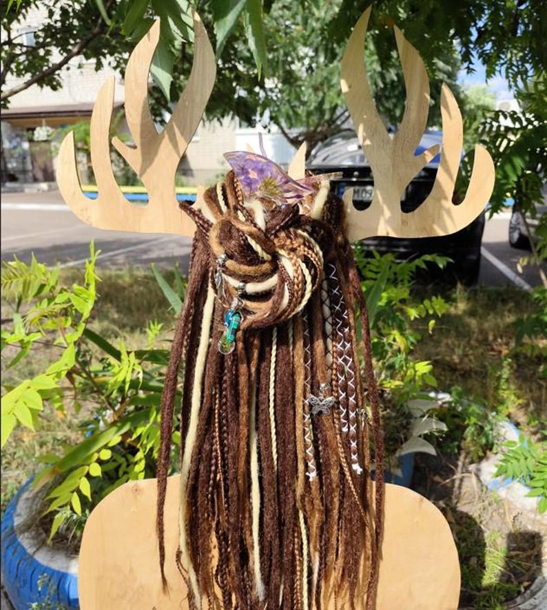 K&ouml;nnte beinhalten: Braune und wei&szlig;e synthetische Dreadlocks mit Perlen und Charms, die daran h&auml;ngen. Die Dreadlocks sind zu einem Knoten gebunden und an einem h&ouml;lzernen Hirschkopf befestigt.