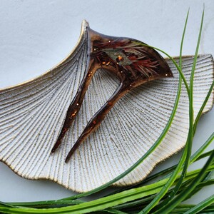 May include: A brown and gold hair clip with a resin design featuring dried flowers and gold glitter. The clip is sitting on a white and gold decorative dish.
