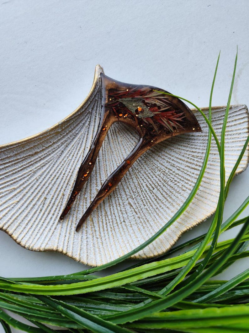 May include: A brown and gold hair clip with a resin design featuring dried flowers and gold glitter. The clip is sitting on a white and gold decorative dish.