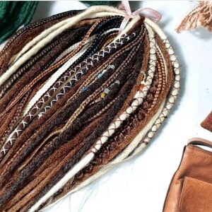 K&ouml;nnte beinhalten: Ein B&uuml;ndel brauner, schwarzer und wei&szlig;er synthetischer Dreadlocks mit wei&szlig;en N&auml;hten. Die Dreadlocks sind mit einem rosa Band zusammengebunden.