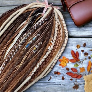K&ouml;nnte beinhalten: Ein B&uuml;ndel brauner, beiger und wei&szlig;er synthetischer Dreadlocks mit wei&szlig;en N&auml;hten und Perlen. Die Dreadlocks sind kreisf&ouml;rmig angeordnet und liegen auf einer Holzoberfl&auml;che.