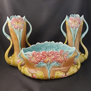 May include: A set of three ceramic vases with floral designs. The vases are in shades of brown, green, and blue, with pink flowers. The vases are in a vintage style.