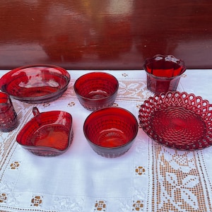 May include: A set of seven red glass bowls and a plate with a decorative pattern. The bowls are various sizes and shapes, and the plate is round with a raised edge.