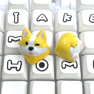 May include: Two yellow and white corgi keycaps with black eyes and a pink tongue. The keycaps are on a white keyboard with black letters.