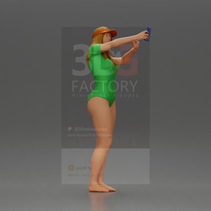 Puede incluir: Una mujer con un traje de baño verde y visera naranja se toma una selfie con un teléfono azul. Está de pie sobre un fondo gris.