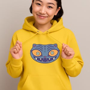 May include: A yellow hoodie with a blue cat face graphic. The cat face has orange eyes, a white-toothed grin, and blue stripes. The hoodie has a drawstring hood and a front pocket.