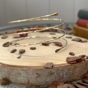 May include: A gold-coloured, hammered-metal arm cuff in a spiral design. The cuff wraps around the arm multiple times. The cuff is displayed on a wooden slice with scattered bark pieces.
