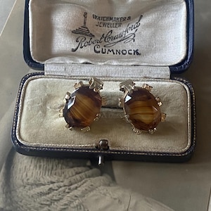 May include: A pair of cufflinks in a black box with a white interior. The cufflinks are made of gold metal and have a brown, marbled stone center. The box has the text "Watchmaker & Jeweller Robert Crawford Cumnock" printed on the inside.