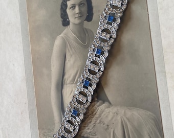 Op de afbeelding: Een zilverkleurige armband met heldere en blauwe strass-steentjes. De armband heeft een herhalend patroon van cirkels en vierkanten, afgebeeld op een vintage zwart-wit foto van een vrouw.