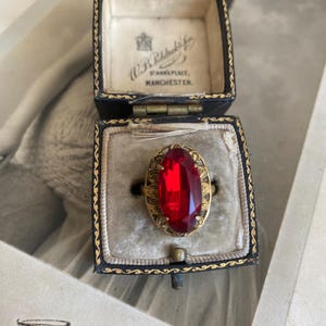 May include: A gold-toned ring featuring a large, oval-cut, deep red faceted stone set within an intricate, filigree-style metal frame. The ring is displayed inside a dark blue, hinged jewelry box with gold trim and a cream-colored velvet interior.