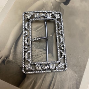 May include: A rectangular, silver-toned belt buckle with a decorative design. The buckle is encrusted with clear, sparkling rhinestones. The design includes floral and crescent moon shapes. The buckle has a central bar and a prong for securing the belt.