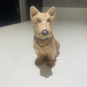 May include: A ceramic figurine of a brown and white dog sitting with its head tilted down. The dog has a black collar and a brown nose.