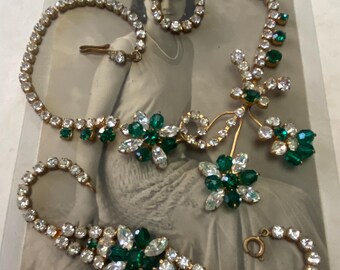 May include: A vintage rhinestone necklace and bracelet set with green and clear stones. The necklace features a floral design with a long, dangling pendant. The set is displayed on a vintage photograph with a signature.