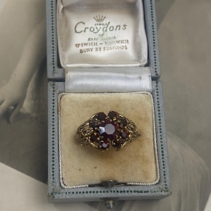 May include: An antique gold-toned ring with a cluster of red gemstones, set in an ornate filigree design. The ring is displayed in a vintage blue jewelry box with the text "Croydons Jewels" visible on the inside lid.