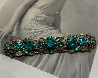 May include: Antique gold-toned barrette featuring emerald green rhinestones. The barrette has an ornate design with floral and leaf accents. The barrette is displayed on a vintage photograph of a woman.