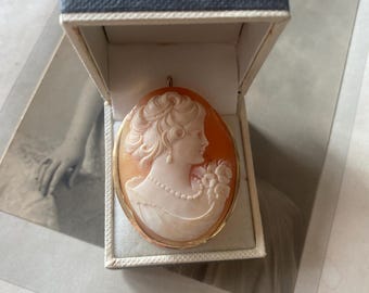 May include: An oval cameo pendant featuring a woman's profile carved in white against an orange background, set in a gold frame. The pendant is displayed in a small, open, blue-gray box, resting on a vintage photograph.