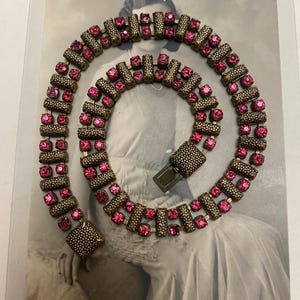 Puede incluir: Un collar vintage de pedrería rosa con un engaste de metal dorado. El collar está hecho de una serie de formas rectangulares unidas, cada una con una fila de pedrería rosa.